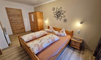 A cozy bedroom with a wooden bed and decorative cushions. The walls are in neutral colors, and there are wooden furniture pieces as well as soft lighting.
