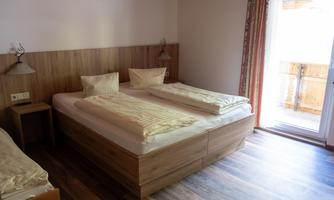 A cozy room with a large double bed and two pillows. There is a balcony, plenty of light, and a modern wooden floor.