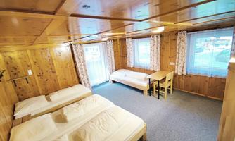 A simple room with wooden walls, a double bed, and a single bed. There is a small table and windows with curtains.