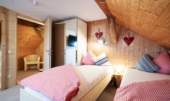 A cozy bedroom with two single beds and wooden walls. The decoration includes cushions with red and white check patterns and heart motifs on the wall.