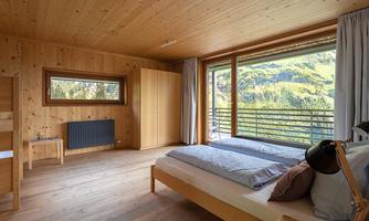 A modern bedroom with wooden walls and large windows. The view overlooks a green mountain landscape.