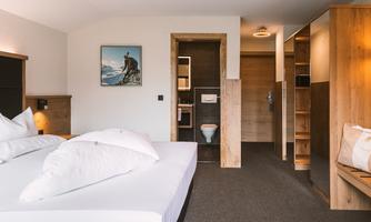 A modern hotel room with a large bed and simple decor. In the background, a bathroom and storage space are visible.
