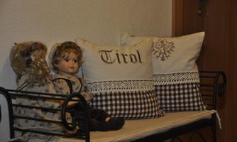 A cozy bench with two cushions that say "Tirol". Next to the cushions sits an older doll with long hair.