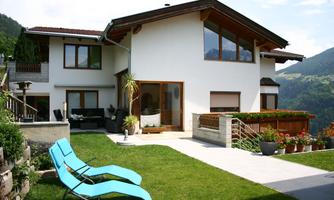 A modern house with large windows and a beautiful garden. In the foreground, there are two loungers on the lawn.