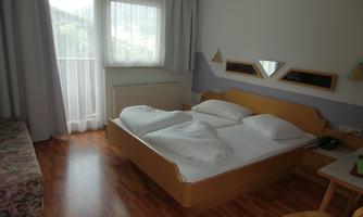 A simple room with a double bed and two pillows. There is a table and an armchair, as well as bright curtains and a balcony.