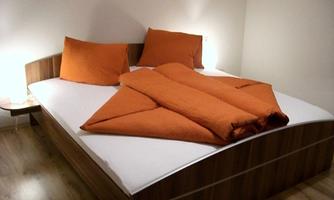 A modern bedroom with a large bed that has orange bedding and pillows. The walls are bright, and the floor is made of wood.