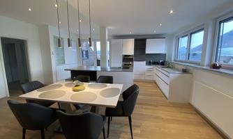 A modern kitchen with a dining area. The space is bright and welcoming, with large windows and an elegant table.