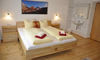 A cozy bedroom with a large bed, equipped with bright blankets and pillows. A picture hangs on the wall, and next to the bed is a sink.