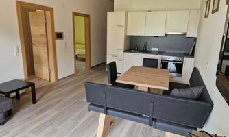 A modern living room with a kitchenette and a dining area. The room has light walls and a warm wooden floor.
