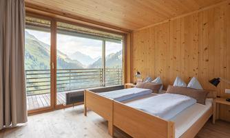 A modern bedroom with wood paneling and large windows. In the background, the mountains and nature are visible.