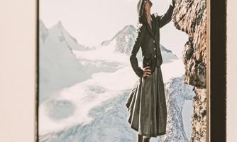 A historic image of a woman in traditional clothing climbing a rock. In the background, snow-capped mountains and a clear sky can be seen.
