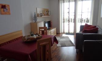 A cozy living space with a dining area and a sofa. Large windows let in plenty of light, and the floor is made of wood.