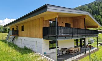 A modern house with wooden cladding and a balcony. It is situated in a green landscape surrounded by mountains.