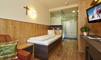 A modern room with wood paneling and a single bed. In the background, there is a television and a shower.