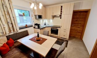 A modern kitchen with a dining area and a large table. The walls are light, and there is comfortable seating with red cushions.