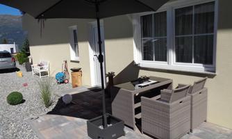 A cozy outdoor area with a table and chairs under a sunshade. The surroundings are well-kept with gravel and a few plants.