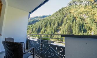 A balcony with a chair and a view of a green mountain landscape. Nature is characterized by trees and hills.