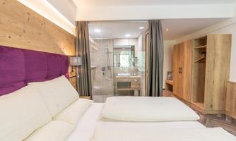A modern room with a double bed and a cozy purple headboard. In the background, you can see a bathroom with transparent walls and wooden details.