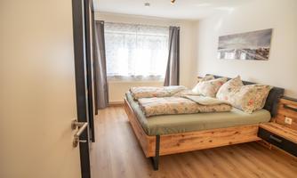 A modern bedroom with a large double bed and romantic floral patterns on the pillows. Large windows let in plenty of natural light and create an inviting atmosphere.