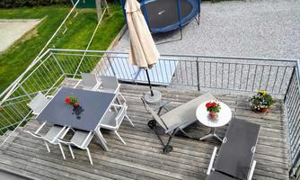 A sunny terrace with a table and chairs, as well as loungers and a sun umbrella. In the background, a trampoline and lawn can be seen.
