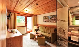 A cozy room with a wooden ceiling and large windows. There is a sofa, a table, and a bathroom area.