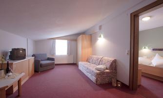 A spacious room with a sofa and an armchair. There is a television, a table, and access to another room.