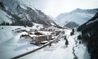 A picturesque mountain landscape in winter with snow-covered hills and impressive peaks. In the foreground, a small village is visible along a road.