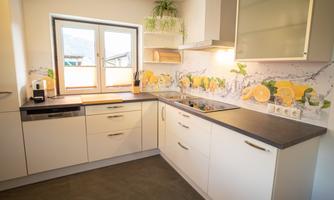 A modern, bright kitchen with white cabinets and a dark gray countertop. On the wall, there are cheerful decorations featuring lemons and plants.