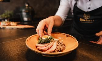 A chef carefully places a serving of pasta in a bowl. The dish consists of pasta, meat, and fresh vegetables.