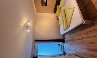 A simple bedroom with a double bed, light pillows, and a large window. The floor is made of wood and there is a closet.