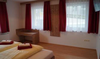 A comfortable bedroom with two beds and red curtains. There is a small workspace and plenty of daylight coming through the windows.