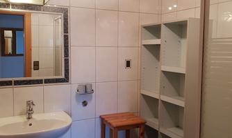 A bathroom with white tiles and a wooden ceiling. There is a sink, a shower stall, and a shelf for storage.