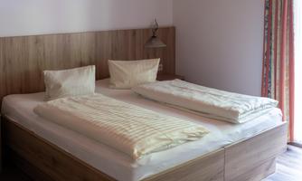 A bright bedroom with a large double bed and two pillows. The wooden floor and the windows with curtains give the room a cozy atmosphere.