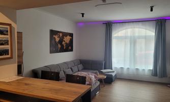 A modern living room with a gray couch and a large window. The walls are decorated, and the lighting is color-coordinated.
