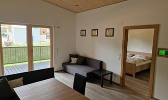 A bright living-dining room with a large window front. There is a table, chairs, and a sofa, as well as access to a bedroom.