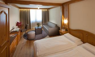 A cozy room with two single beds and wooden paneling. There is a sofa, a table, and large windows that let in plenty of light.