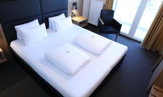 A modern bedroom with a large double bed and two pillows. In the background, there is a chair and large windows with curtains.