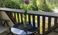 A cozy balcony with a round table and a blooming vase. In the background, green hills and trees can be seen.