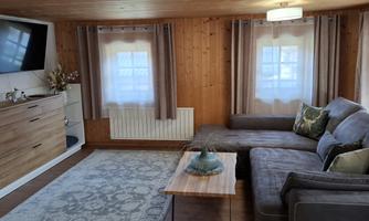 A cozy living room with a gray couch and a wooden table. The room features a television and large windows with curtains.