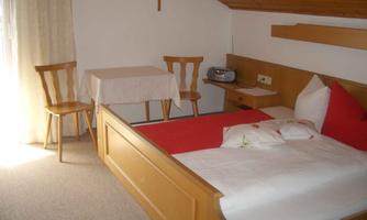 A simple bedroom with a double bed, a table, and two chairs. There is a wooden wardrobe and a window with curtains.