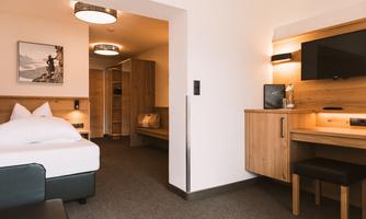 A modern hotel room with a large bed and a cozy seating area. The walls are in warm wooden tones, and there is a workspace with a TV.
