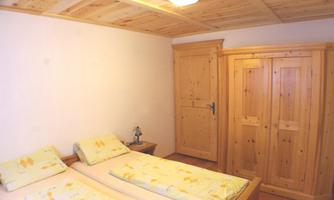 A simple bedroom with two bed frames and wooden furniture. The room has a natural wooden ceiling and a wardrobe.