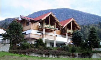 A large, modern house with a red roof and wooden cladding. In the background, mountains and a green garden can be seen.