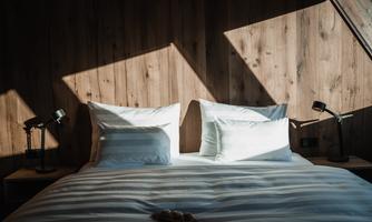 A cozy bedroom with a large bed and white pillows. Soft light comes through the window and illuminates the wooden wall.