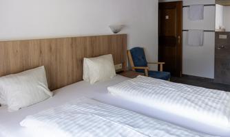 A simple bedroom with two single beds and a wooden panel wall. The room is bright and furnished in a modern style, with a blue armchair in the corner.