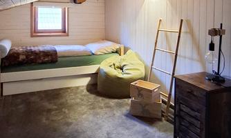 A cozy bedroom with a single bed, a bean bag, and wooden furniture. The walls are made of wood, and a window provides daylight.