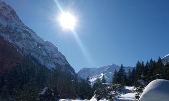 A clear winter landscape with a snow-covered path and high mountains. The sun shines brightly in the blue sky.