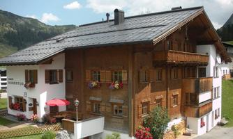 A charming alpine house with wooden cladding and a modern extension. Colorful flowers adorn the windows, and the garden is well-maintained.