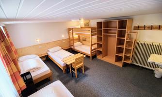 A simple room with several beds and a table. There is also a wardrobe and space for personal items.