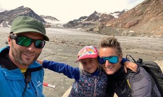 A family poses smiling in front of an impressive mountain landscape. Behind them, glaciers and tall mountains can be seen.